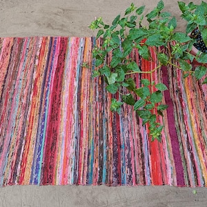 Könnte beinhalten: Ein rechteckiger, mehrfarbiger Flickenteppich mit vertikalen Streifen in Rot-, Rosa-, Orange- und Brauntönen. Der Teppich hat einen Fransenrand und liegt auf einer neutralen Oberfläche. Eine Topfpflanze mit grünen Blättern befindet sich in der oberen rechten Ecke.