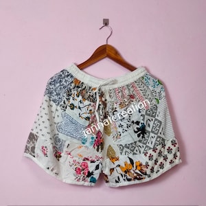 May include: Patchwork shorts in white with a drawstring waist. The shorts display floral and geometric patterns in pink, blue, yellow, and grey. They are suspended from a wooden hanger.