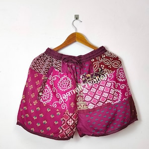 May include: Patchwork shorts in shades of magenta, burgundy, and teal, hanging on a wooden hanger. The shorts feature a drawstring waist and a variety of patterned fabric patches, including geometric and floral designs. The text "jannalcreation" is visible on the shorts.