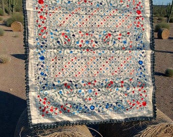 Può includere: Un tessuto bianco con ricami intricati in motivi floreali rossi e blu. I bordi sono rifiniti con un volant blu scuro. Il tessuto è esposto all'aperto, appoggiato su una balla di fieno, con un paesaggio desertico sullo sfondo.