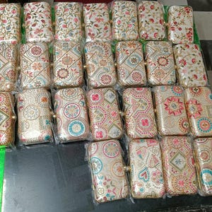 Puede incluir: Una colección de pequeños bolsos de mano rectangulares blancos con intrincados bordados florales y geométricos en tonos de rosa, azul y verde. Los bolsos tienen cierres de metal dorado y están dispuestos en filas.