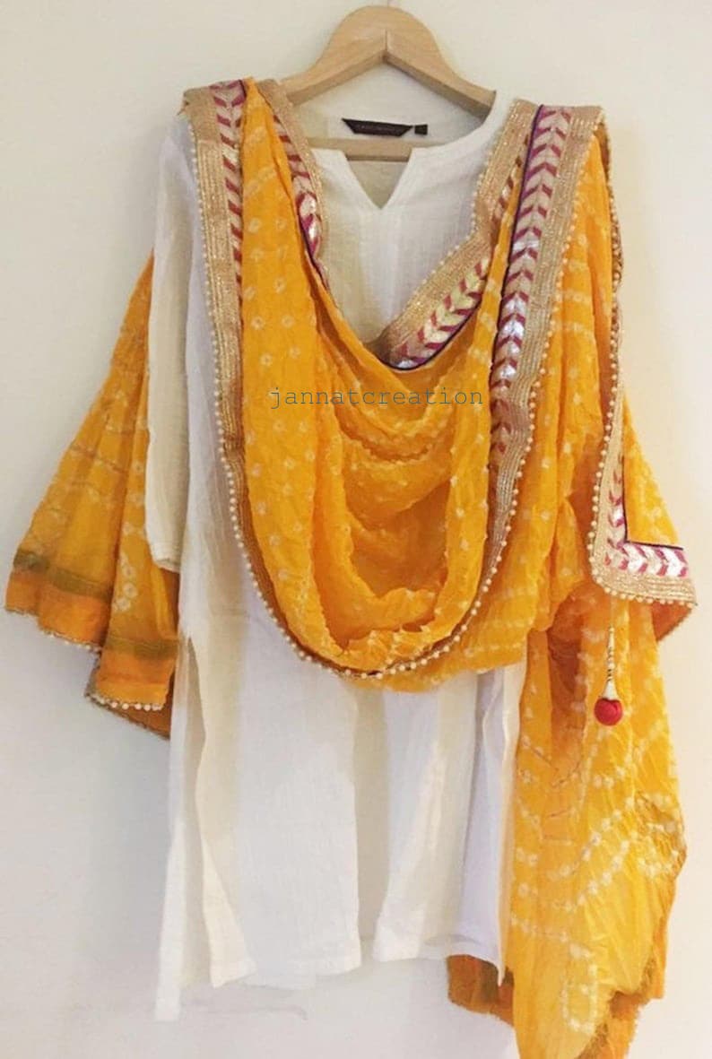 May include: White tunic and a bright yellow dupatta with a dotted pattern and decorative trim. The dupatta is draped over the tunic, creating a layered effect. The outfit is hanging on a wooden hanger.