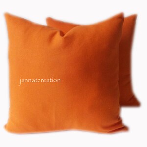 May include: Two square orange cushions. The cushions are a solid, vibrant orange colour and appear to be made of a soft fabric. The text "jannatcreation" is printed on the front of the cushion.