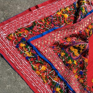 May include: A vibrant red textile with intricate embroidery. The design features white, yellow, green, and blue floral patterns, accented with small, round embellishments. The edges are trimmed with white stitching and a blue border.