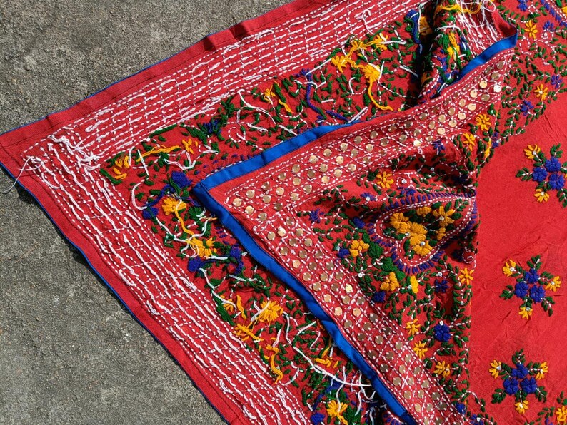 May include: A vibrant red textile with intricate embroidery. The design features white, yellow, green, and blue floral patterns, accented with small, round embellishments. The edges are trimmed with white stitching and a blue border.