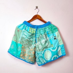 May include: Patchwork shorts in shades of teal, turquoise, and green, with a blue drawstring waistband and trim. The shorts feature a variety of patterned fabric patches and a small embroidered design. The shorts are hanging on a wooden hanger.