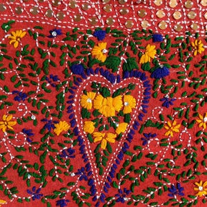 May include: Close-up of a red textile with intricate embroidery. The design features yellow and blue flowers, green leaves, and a heart-shaped floral arrangement. The top edge has a blue border and decorative sequins.