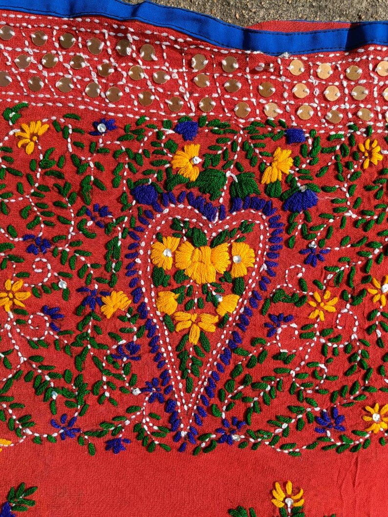 May include: Close-up of a red textile with intricate embroidery. The design features yellow and blue flowers, green leaves, and a heart-shaped floral arrangement. The top edge has a blue border and decorative sequins.