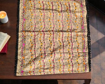 May include: Beige rectangular textile with a floral pattern in pink, yellow, and orange. The edges are trimmed with black fringe. The textile is placed on a dark brown wooden table.