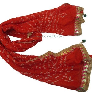 May include: A vibrant red scarf with a dotted pattern and gold trim. The scarf has a decorative border with pink accents and green tassels. The fabric appears to be lightweight and flowing, suitable for draping.