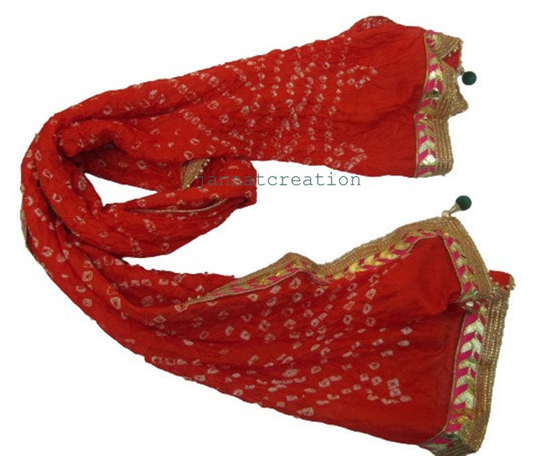 May include: A vibrant red scarf with a dotted pattern and gold trim. The scarf has a decorative border with pink accents and green tassels. The fabric appears to be lightweight and flowing, suitable for draping.