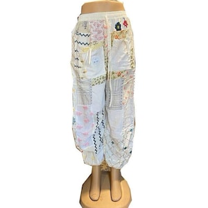 Op de afbeelding: Witte patchworkbroek met diverse patronen, waaronder bloemen, strepen en zigzagontwerpen. De broek heeft een taille met trekkoord en is bij de enkels gerimpeld. De stof lijkt lichtgewicht, geschikt voor warm weer.