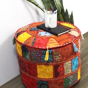 May include: A round, cylindrical ottoman featuring a patchwork design of vibrant, multi-colored fabric squares in shades of red, orange, yellow, and blue, accented with decorative tassels along the top edge and intricate embroidery throughout.