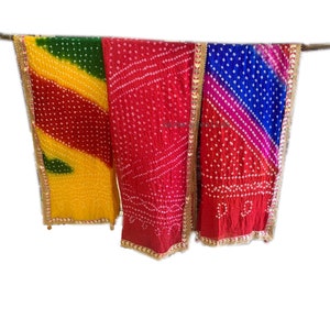 May include: Three colorful, patterned scarves hanging against a white background. The scarves are in shades of yellow, green, red, and blue, with white dot patterns and gold trim. The fabric appears to be lightweight.
