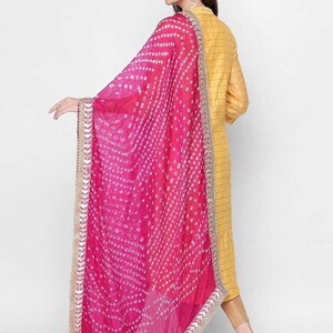 May include: A vibrant pink dupatta with a white dotted pattern and a gold border. The dupatta is draped over a yellow outfit. The fabric appears lightweight and flowing, with a gradient of pink hues.