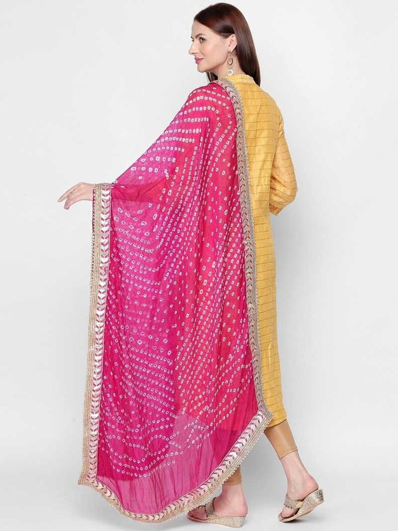 May include: A vibrant pink dupatta with a white dotted pattern and a gold border. The dupatta is draped over a yellow outfit. The fabric appears lightweight and flowing, with a gradient of pink hues.