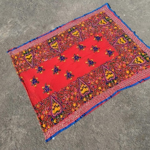 May include: A vibrant red textile with intricate embroidery. The square fabric features a central panel with floral and star-like patterns, surrounded by paisley and floral designs in yellow, blue, and orange. The edges are trimmed with blue and silver.