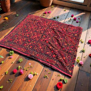 Peut inclure: Un tapis rectangulaire à motifs rouge foncé et noir avec une bordure rouge. Le tapis est posé sur un plancher en bois, avec des fleurs et de la verdure éparpillées autour. Le motif du tapis présente des motifs floraux et géométriques.