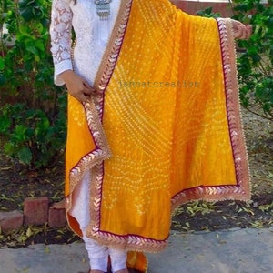 May include: A woman's white embroidered tunic and leggings paired with a vibrant yellow dupatta. The dupatta features a detailed pattern and a decorative border with gold and purple accents. The text "jannatcreation" is visible on the dupatta.