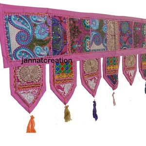 May include: A pink textile wall hanging with embroidered paisley and floral patterns. The wall hanging has multiple rectangular and triangular panels with colorful designs and tassels. The text "jannatcreation" is visible on the wall hanging.