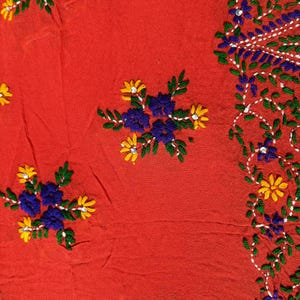 May include: Close-up of a red fabric with embroidered floral patterns. The embroidery features blue, yellow, and green flowers and leaves. The design includes clusters of flowers and a border of vines.