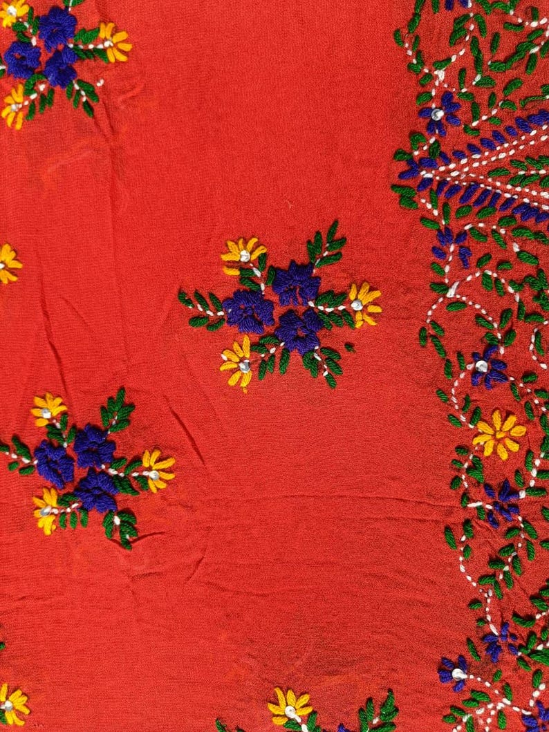 May include: Close-up of a red fabric with embroidered floral patterns. The embroidery features blue, yellow, and green flowers and leaves. The design includes clusters of flowers and a border of vines.