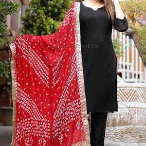 May include: Black kurta and pants ensemble paired with a vibrant red dupatta. The dupatta features a white geometric pattern and gold trim. The outfit is a traditional Indian style, suitable for festive occasions.