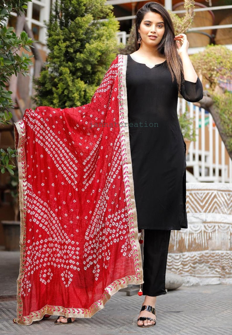May include: Black kurta and pants ensemble paired with a vibrant red dupatta. The dupatta features a white geometric pattern and gold trim. The outfit is a traditional Indian style, suitable for festive occasions.