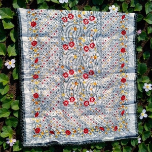 May include: A white textile with intricate embroidery featuring red, yellow, and green floral patterns. The design includes a diamond grid and a border with a dark trim. The textile is displayed on a bed of green leaves and white flowers.