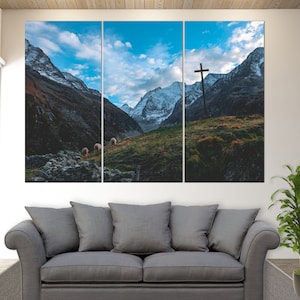Op de afbeelding: Een driedelig canvasprint met een schilderachtig berglandschap met een houten kruis gesilhouetteerd tegen een blauwe lucht. De bergen zijn met sneeuw bedekt en op de voorgrond is een grasveld met schapen te zien.