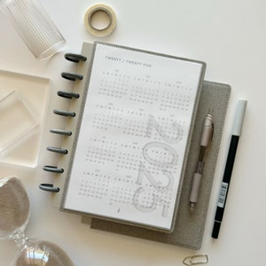 May include: A gray and white planner with a clear plastic cover. The planner has a calendar for the year 2024 with the numbers 2024 printed in a gray, semi-transparent font. The planner is open to the month of January.