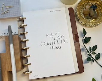 May include: A brown leather planner with gold-colored rings, open to a page with the text "beginning is EASY CONTINUING is hard." Other items include a gold binder clip, pens, and decorative stones.