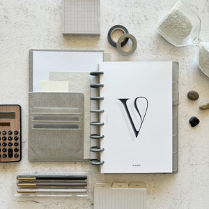 May include: A gray and white planner with a black letter V on the cover. The planner has a ring binding system and a pocket on the front cover. The planner is on a white surface with other office supplies.
