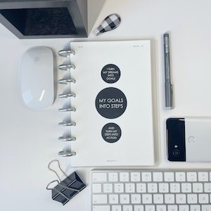 May include: A white planner with black text and circles. The planner has a silver coil binding and the text on the cover reads "I turn my dreams into goals", "My goals into steps", and "And turn my steps into action".