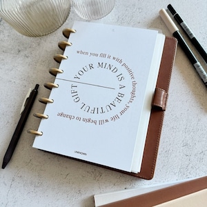 May include: A white planner with a brown leather cover. The planner has a gold ring binding and a quote that reads "When you fill it with positive thoughts, your mind is a beautiful gift. Life will begin to change."