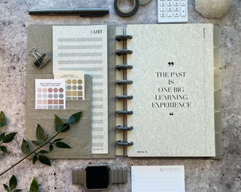 May include: A gray and white planner with a quote that reads "The past is one big learning experience". The planner has a ring binding system and a gray linen cover. The planner is open to a page with a list of events.