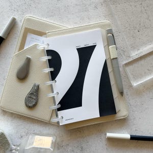 May include: A disc-bound planner with a cream-colored textured cover, featuring a white and black insert with the large text 27 and the words TWENTY TWENTY SEVEN. Includes a grey pen, two small grey clips, and a small card labeled JOT THIS.