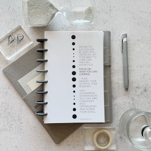 May include: A white planner with black text that reads "The path to success may be filled with obstacles, but your ability to overcome them lies within you. Focus on what you can control - your mindset, your actions, your resilience. With unwavering determination, you can turn challenges into opportunities and achieve your goals. AndPaperCo."