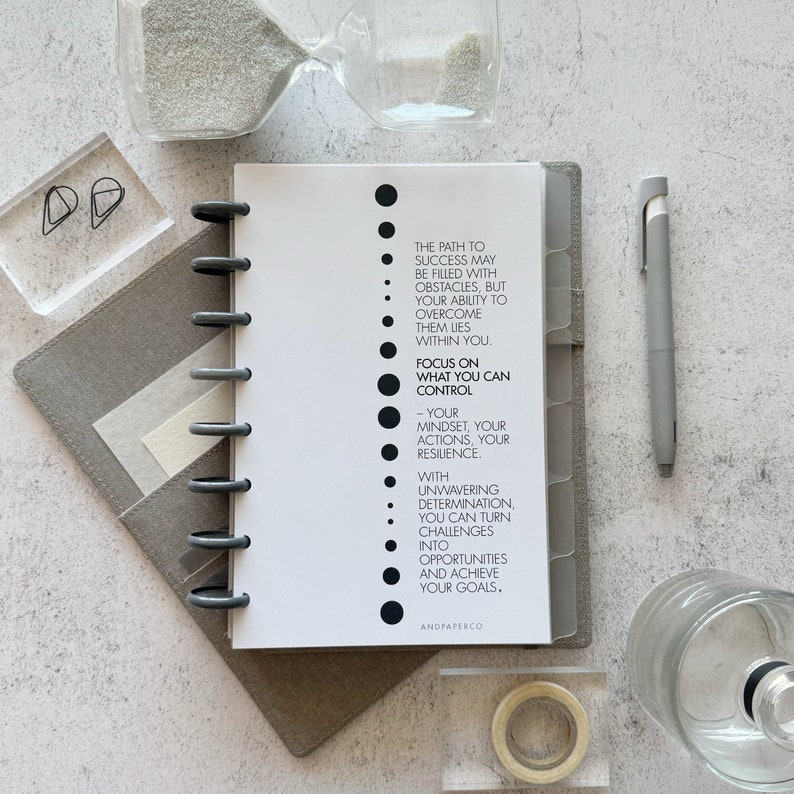 May include: A white planner with black text that reads "The path to success may be filled with obstacles, but your ability to overcome them lies within you. Focus on what you can control - your mindset, your actions, your resilience. With unwavering determination, you can turn challenges into opportunities and achieve your goals. AndPaperCo."