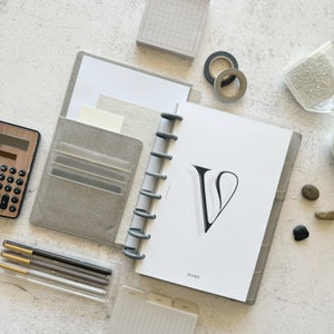 May include: A gray and white planner with a black letter V on the cover. The planner has a ring binding system and a clear plastic pocket on the front cover. The planner is open to a page with the word "NAME" printed at the bottom.