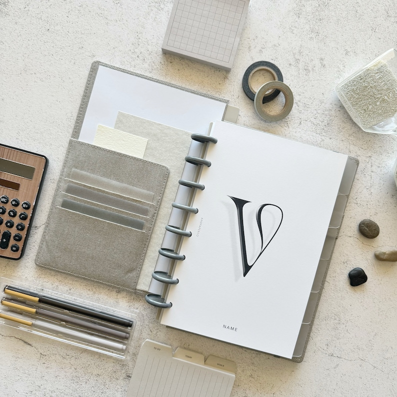May include: A gray and white planner with a black letter V on the cover. The planner has a ring binding system and a clear plastic pocket on the front cover. The planner is open to a page with the word "NAME" printed at the bottom.