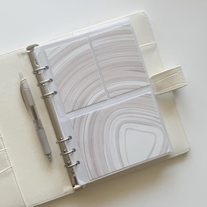 May include: An open, off-white leather binder with silver metal rings and a matching pen. The binder contains clear plastic sleeves holding patterned cards with swirling gray and white designs. The binder is closed with a strap.