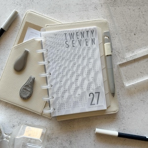 May include: A cream-colored disc-bound planner with a translucent cover featuring the text TWENTY SEVEN and a calendar grid. The planner includes a grey pen, two small grey charms, and a grey marker, all resting on a textured surface.