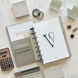 May include: A gray and white planner with a large letter V on the cover. The planner has a ring binding system and a pocket on the front cover. The planner is open to a page with the word "NAME" at the bottom.