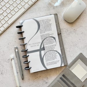 May include: A gray planner with a spiral binding, open to a page with the text "TWO THOUSAND TWENTY SIX". The planner is next to a white keyboard, a white mouse, and two gray pens. A gray wallet is also visible.