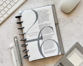 May include: A gray planner with a spiral binding, open to a page with the text "TWO THOUSAND TWENTY SIX". The planner is next to a white keyboard, a white mouse, and two gray pens. A gray wallet is also visible.