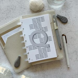 May include: A beige leather-textured planner with white pages featuring the repeated text Quincie Brianne Garcia. A large silver letter Q sits on the page. A gray pen is held in the planner loop, and a white pen lies next to the planner.