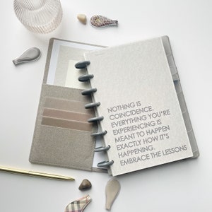 May include: A gray and white notebook with a motivational quote: "Nothing is coincidence. Everything you're experiencing is meant to happen exactly how it's happening. Embrace the lessons."