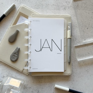 May include: A white disc-bound planner with a cream-colored textured cover. The cover page features the text JAN, 2025 / QUARTER 01, CHAPTER 01, and ANDPAPERCO. A gray pen is attached to the side, accompanied by two small gray clips and two pens.