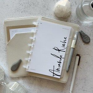 May include: A cream-colored textured planner cover with a white disc-bound notebook insert. The cover page features the text Amanda Riahe in black script, with A/R and San Francisco printed above. Two pens, one gray and one white, are included.